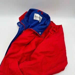 Puede incluir: Chaqueta roja y azul con cierre de cremallera y un bolsillo en el lado izquierdo. La chaqueta tiene una etiqueta que dice "Saxton Hall" hecha en los Estados Unidos.