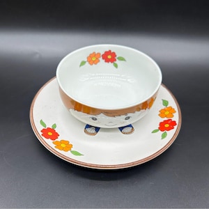 Puede incluir: Un juego vintage de cuenco y platillo de cerámica. El cuenco y el platillo son blancos con un patrón moteado y un borde marrón. El cuenco presenta un diseño de niño de dibujos animados con pelo naranja y zapatos azules. El platillo está decorado con flores naranjas y rojas.