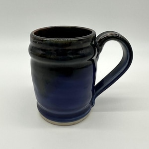 May include: A handmade ceramic mug with a dark blue and black glaze. The mug has a rounded body, a sturdy handle, and a light-colored base. The interior is dark brown. This mug is perfect for coffee, tea, or any hot beverage.