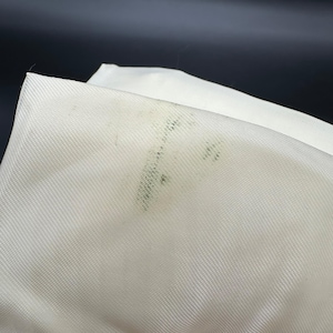 May include: Close-up of a folded white fabric with a visible diagonal weave. The fabric has several dark green spots on the surface, likely from mold or mildew. The background is a dark, solid color.