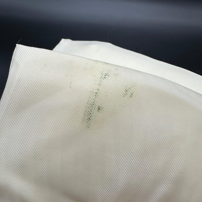 May include: Close-up of a folded white fabric with a visible diagonal weave. The fabric has several dark green spots on the surface, likely from mold or mildew. The background is a dark, solid color.