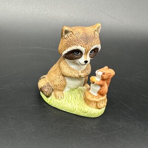 May include: A ceramic figurine featuring a raccoon and a squirrel. The raccoon is brown and white with a black mask, sitting on a patch of green grass. The squirrel is orange and white, holding an acorn. The figurine is on a green base.