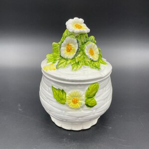 May include: White ceramic jar with a lid decorated with white flowers and green leaves. The jar has a woven pattern and is accented with yellow flower centers.