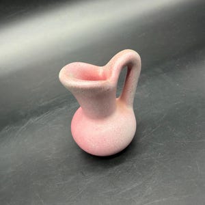 Puede incluir: Una pequeña jarra de cerámica con cuerpo redondeado y asa curva. La superficie presenta un acabado mate y moteado que transiciona desde un rosa suave en la base hasta un tono rosa pálido más claro cerca del pico vertedor asimétrico.