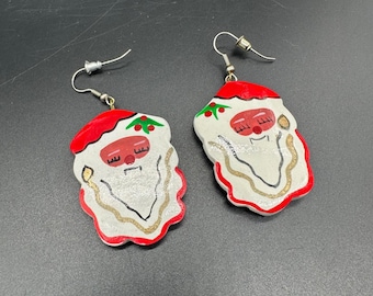 May include: A pair of Santa Claus face earrings. The earrings feature a red hat, white face, and a gold beard. The Santa faces have closed eyes and a red mouth. The earrings are made of a painted material and have silver hooks.