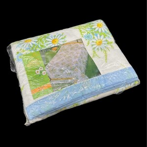 May include: A packaged white bedspread with a daisy floral pattern in yellow, green, and blue. The bedspread is wrapped in clear plastic. The packaging includes a picture of the bedspread on a bed.