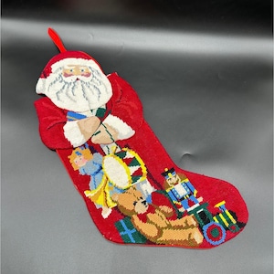 May include: A red Christmas stocking with a Santa Claus design at the top. The stocking features a needlepoint design of a drum, teddy bear, toy soldier, and other toys. A red loop is attached to the top.