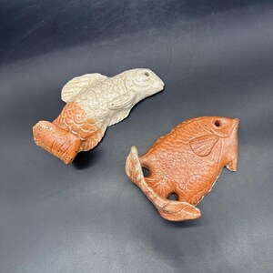 以下が含まれることがあります： 2つのセラミック製の魚の彫刻。1つはオフホワイトにオレンジ色のアクセント、もう1つはオレンジ色です。どちらも詳細な鱗とひれがあります。魚は装飾品である可能性があります。