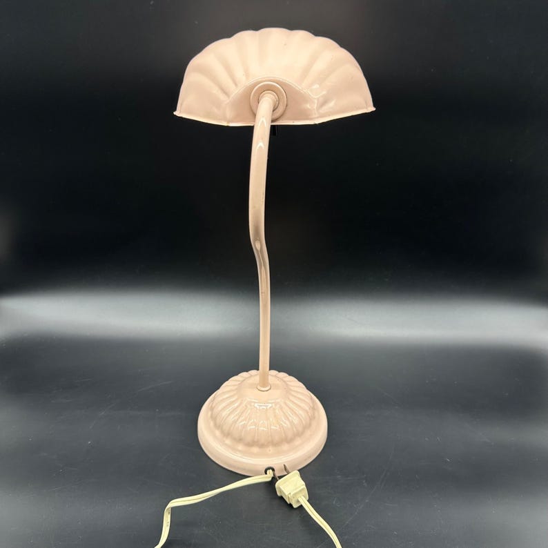 K&ouml;nnte beinhalten: Eine Schreibtischlampe im Vintage-Stil in einem weichen, ged&auml;mpften Rosa. Die Lampe hat einen gewellten Schirm, einen gebogenen Hals und einen dekorativen Sockel. Das Kabel und der Stecker sind sichtbar. Diese Lampe ist eine dekorative Leuchte.
