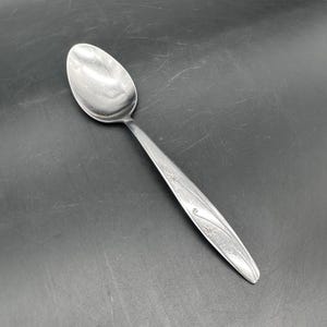 Puede incluir: Una cuchara de acero inoxidable con un cuenco redondeado y un mango largo y delgado. El mango presenta un diseño decorativo grabado con motivos de estrellas y espirales. La cuchara está diseñada para comer y servir.