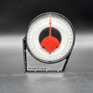 May include: A black and clear magnetic base protractor with a red indicator. The protractor has degree markings from 0 to 90. The text "MAGNETIC BASE" is printed on the clear base. The protractor is used for measuring angles.