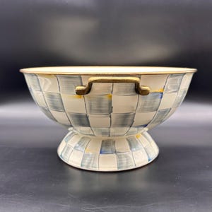 Op de afbeelding: Een kom op voet met een handgeschilderd blauw en wit ruitpatroon over de buitenkant en de basis. De kom heeft een brede, open bovenrand en twee goudkleurige metalen handvatten aan de zijkanten, die een contrast vormen met het patroon.