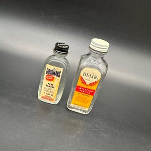 Puede incluir: Dos botellas de vidrio de saborizante. La botella de la izquierda está etiquetada como "Goodman's Pure Almond Flavoring" y contiene 1 onza líquida. La botella de la derecha está etiquetada como "Blair Imitation Vanilla Flavoring" y contiene 29,5 ml.