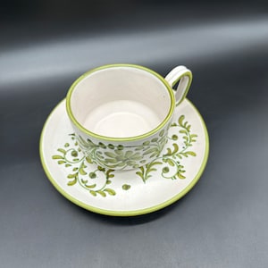 Może przedstawiać: Biały ceramiczny kubek do herbaty i spodek z zielonym wzorem kwiatowym. Kubek ma zaokrąglony kształt i uchwyt. Spodek ma falisty brzeg.