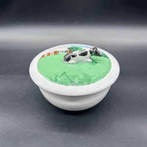 May include: A white ceramic butter dish with a lid featuring a painted scene. The lid has a green field with a black and white cow figurine. A fence and blue sky are also painted on the lid.