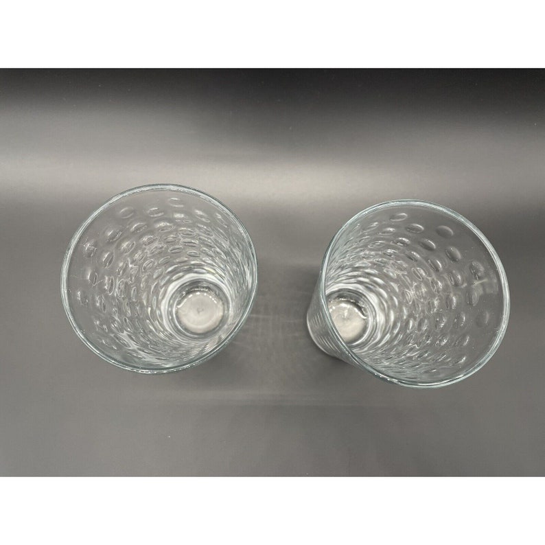 May include: Two clear glass tumblers with a textured, bubbled design. The glasses are empty and sitting on a gray surface.