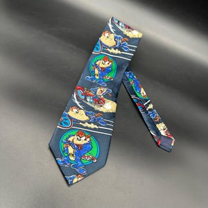 May include: A navy blue necktie featuring a repeating pattern of the Tasmanian Devil cartoon character in various action poses. The tie has white stripes and circular designs with the character in a blue and red outfit.