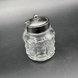 May include: A clear glass jar with a silver-colored lid and a small handle. The jar has a textured, starburst-like pattern. The lid has a small, round knob on top. The jar is likely for storing condiments or other small items.