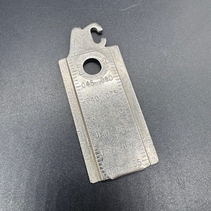 May include: A silver-colored spark plug gap gauge with a hook, a hole, and measurement markings. The gauge has numbers indicating measurements in inches, such as .020, .025, .030, .035, .040, .045, .050, .055, .065, .075, and .080.