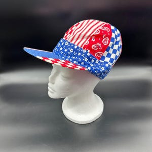 May include: A red, white and blue patchwork baseball cap with a patterned brim. The cap is made of different fabrics, including a paisley print, a floral print and a checkered print.