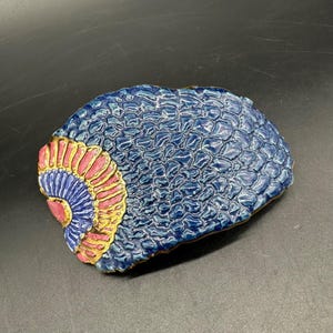 May include: A ceramic decorative dish with a textured blue surface and a fan-shaped design in pink, yellow, and blue. The dish has a unique, organic shape and is likely handmade.