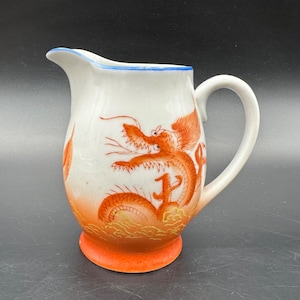 May include: White ceramic pitcher with a dragon design. The pitcher features a curved handle and a spout with a blue rim. The dragon is orange and red, with orange accents at the base.