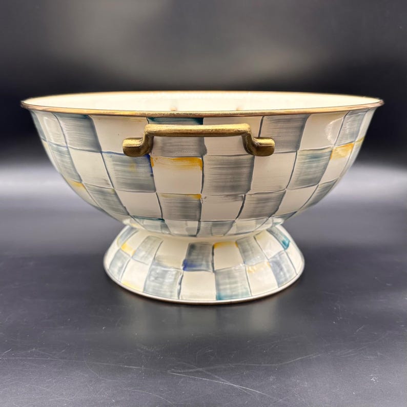 Op de afbeelding: Een kom op voet met een handgeschilderd ruitpatroon in blauw-, grijs- en geeltinten op een gebroken witte achtergrond. De kom heeft een metallic goudkleurige rand en twee bijpassende goudkleurige handvatten aan weerszijden van de buitenkant.