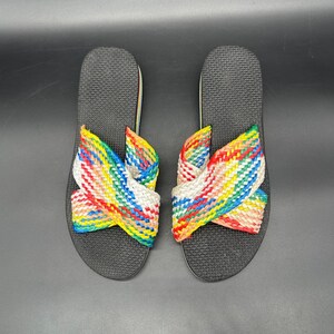 May include: A pair of black flip-flops with woven rainbow straps. The straps are a mix of red, yellow, green, blue, and white, creating a colorful design. The flip-flops are set against a dark background.