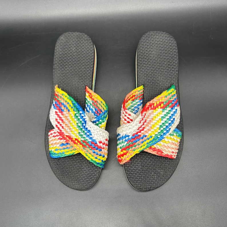 May include: A pair of black flip-flops with woven rainbow straps. The straps are a mix of red, yellow, green, blue, and white, creating a colorful design. The flip-flops are set against a dark background.