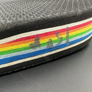 May include: Close-up of a black shoe sole with a rainbow-striped edge. The sole has a textured, wavy pattern. The rainbow stripes include red, orange, yellow, green, blue, and purple. There are faded markings on the rainbow stripes.