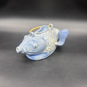 以下が含まれることがあります： 青と白の釉薬を施した、魚の形をした陶器のティーポット。魚には注ぎ口、取っ手、蓋が付いています。ティーポットは花の模様と黄色のコードで飾られています。
