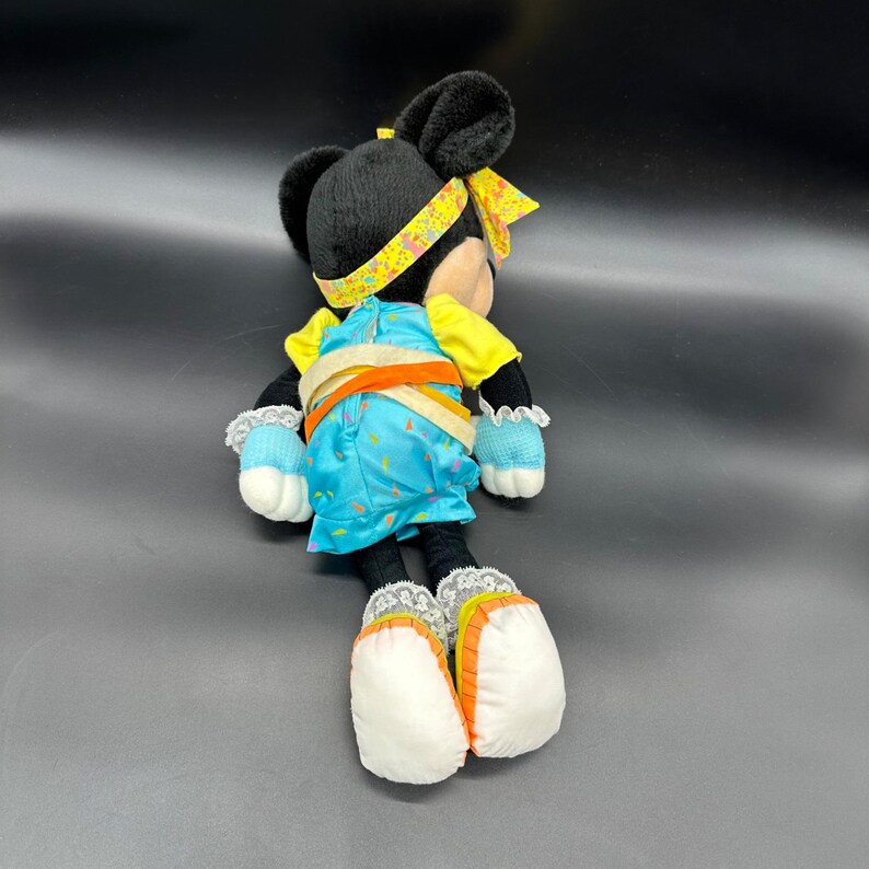 Peut inclure: Un jouet en peluche de Minnie Mouse portant une robe bleue et jaune avec un bandeau blanc. Le jouet est allong&eacute; sur le dos, les jambes tendues.