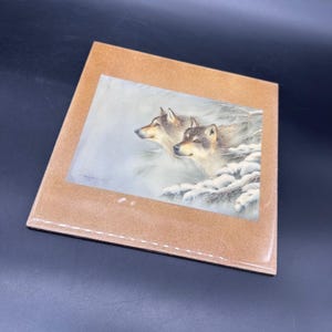 May include: A framed print featuring two wolves in a snowy landscape. The artwork depicts the wolves with detailed fur and a serene expression, set against a backdrop of snow-covered branches and a soft, muted color palette. The frame is a light brown color.