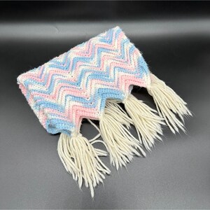May include: A crocheted scarf featuring a repeating chevron pattern in light blue, soft pink, and white yarn. The edges of the scarf are finished with long, thick, cream-colored fringe tassels that hang from the bottom points of the chevron design.