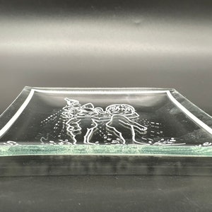 May include: A clear glass serving tray with a raised etched design of two children playing. The tray has a slightly rounded edge.