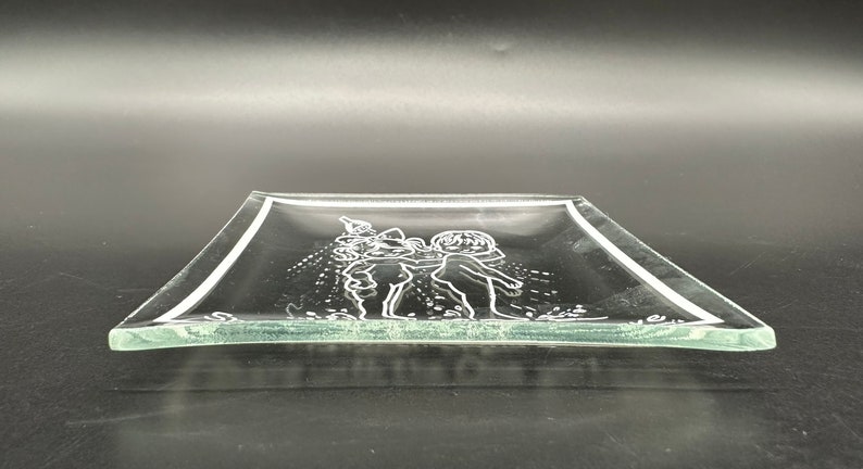May include: A clear glass serving tray with a raised etched design of two children playing. The tray has a slightly rounded edge.