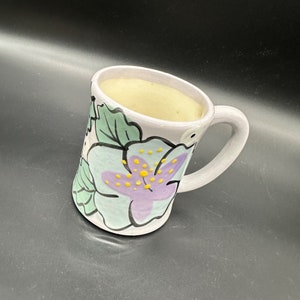 May include: A white ceramic mug with a floral design. The mug features a purple flower with yellow dots and green leaves. The mug is filled with a light brown liquid.