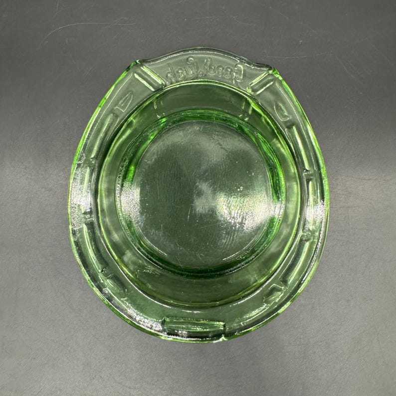 May include: A vintage green glass ashtray with a circular design. The ashtray has a rounded shape with a slightly raised rim and a central depression. The text "Candy Dish" is embossed on the rim.