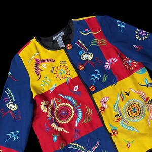May include: A colorful jacket with a patchwork design. The jacket features embroidered floral and abstract patterns in yellow, red, and blue. It has a black collar and is fastened with round, orange buttons. The label reads "Indigo Moon".