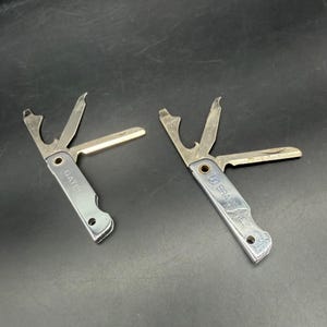 Puede incluir: Dos herramientas múltiples de metal plateado con componentes plegables. La herramienta de la izquierda tiene grabado el nombre GATES. La herramienta de la derecha presenta el texto BRA INC. Cada una incluye un abridor, una hoja y un borde plano.