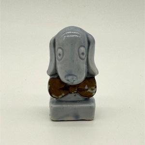 May include: A light blue ceramic dog figurine with a brown bow tie. The dog has large, round eyes and floppy ears. The figurine is standing on a small, rectangular base. A decorative item.