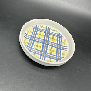 May include: A small, off-white ceramic dish with a plaid pattern in yellow, blue, and brown. The dish has a slightly raised edge and a smooth, glossy finish. The plaid design covers the interior base of the dish.