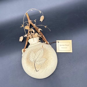 May include: A beige stoneware vase with a leaf design and dried plant stems. The vase has a round shape with a narrow neck and a tag that reads "Jimmie Floyd Stoneware."
