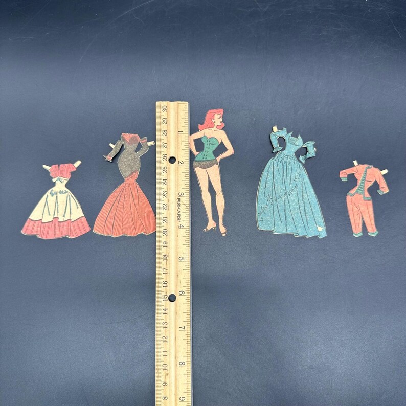 Puede incluir: Una colecci&oacute;n de mu&ntilde;ecas de papel y trajes vintage, que incluyen vestidos y una figura. Se coloca una regla de madera junto a las mu&ntilde;ecas. La regla mide aproximadamente 30 cm.