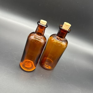 Puede incluir: Dos botellas de vidrio ámbar con tapones de corcho. Las botellas están vacías y tienen un aspecto vintage.