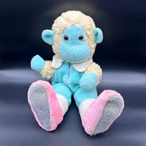 Peut inclure: Un singe en peluche avec un corps bleu clair, une t&ecirc;te cr&egrave;me et des pieds roses et gris. Le singe porte une combinaison bleu clair avec un col blanc et une fermeture &eacute;clair. Le jouet a des yeux rouges et est assis.