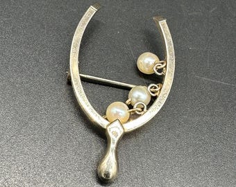 Puede incluir: Un broche de plata en forma de herradura con una cadena de perlas falsas colgantes.
