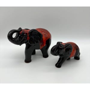 May include: Two ceramic elephant figurines. The larger elephant has a red and black glaze, while the smaller one has a similar design. The elephants are standing on a white surface.