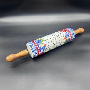 Puede incluir: Un rodillo de cerámica con un estampado floral en azul, rojo y amarillo. El rodillo tiene mangos de madera y una base blanca con detalles de estrellas azules. El diseño floral incluye un borde rojo.