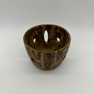 May include: A small, brown and green ceramic bowl with a unique design. The bowl has several holes around the rim and a textured surface.